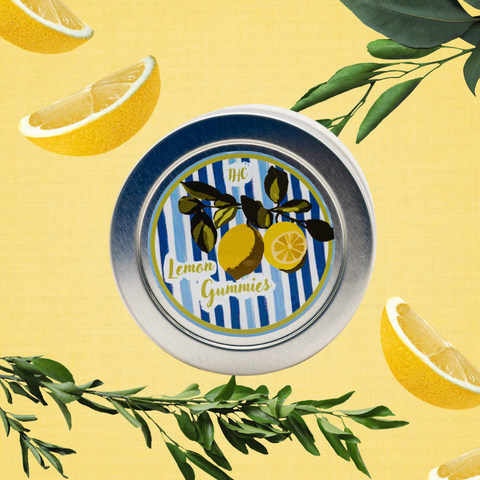 A round tin of lemon gummies with a blue and white striped label featuring yellow lemons and green leaves, set against a yellow background decorated with lemon slices and green foliage.