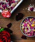 An 8 oz tin filled with bath salts, rose petals, chamomile flowers, and lavender from Appalachian Standard's CBD Botanical Dream Bath Soak on a piece of wood surrounded by flowers