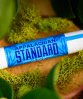 A stick of Appalachian Standard's Vanilla Spearmint CBD Lip Balm on a piece of wood surrounded by plants