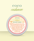 Appalachian Standard Coco Cashmere CBD Body Cream tin, 188mg, 1.5 oz, displayed on a soft yellow background. The round tin features a cream and coral label with a daisy pattern.