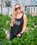 Woman standing among plants in a greenhouse wearing a black tank top with text.