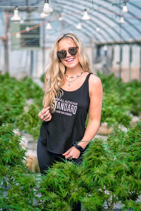 Woman standing among plants in a greenhouse wearing a black tank top with text.