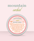Appalachian Standard Mountain Orchid CBD Body Cream tin, 188mg, 1.5 oz, displayed on a soft pink background. The round tin features a blush and coral label with a daisy pattern.