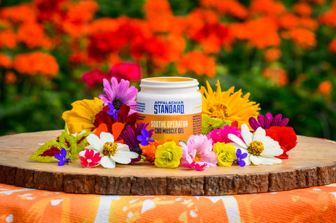 Appalachian Standard Soothe Operator CBD Muscle Gel container, with the lid off, displayed on natural wood round with an artistic arrangement of colorful garden flowers - yellow marigolds, purple asters, white cosmos, pink carnations, and red blooms.