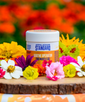 Appalachian Standard Soothe Operator CBD Muscle Gel jar with white lid positioned on a rustic wood slice, surrounded by vibrant wildflowers including yellow zinnias, purple cosmos, white daisies, and pink blooms against a soft-focus orange and green floral background.